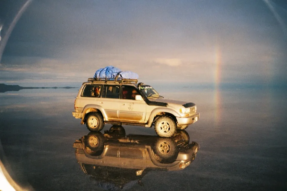 A loaded 4WD reflected in the flooded Salar de Uyuni at dusk, a faint rainbow behind