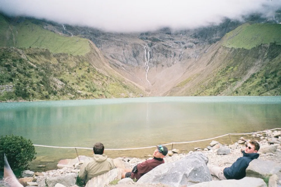 Three hikers resting by the turquoise Humantay Lake below a waterfall